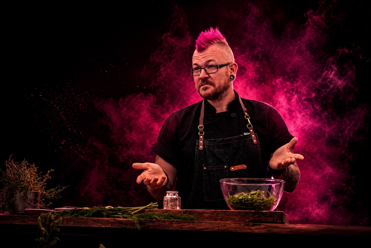 Punk Chef behind a kitchen worktop, surrounded by vivid pink smoke with his hands open
